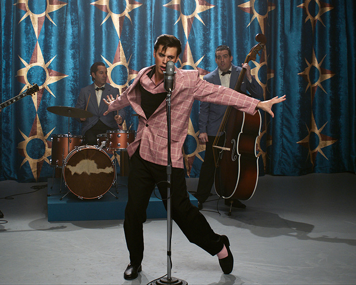 Austin Butler performing energetically in front of a vintage microphone during a band photoshoot with a retro backdrop. Austin Butler performing energetically in front of a vintage microphone during a band photoshoot with a retro backdrop.