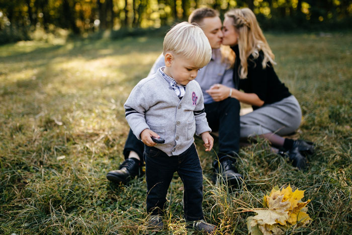 Young boy standing in a grassy field while parents sit behind, highlighting parents obsessed with having kids and older son babysitting. Young boy standing in a grassy field while parents sit behind, highlighting parents obsessed with having kids and older son babysitting.