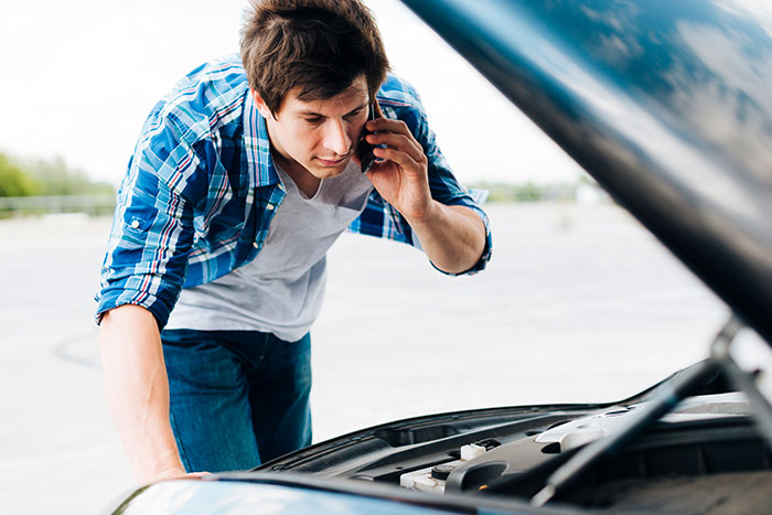 Young man experiencing reverse culture shocks after moving back home, checking a broken car engine while on the phone.