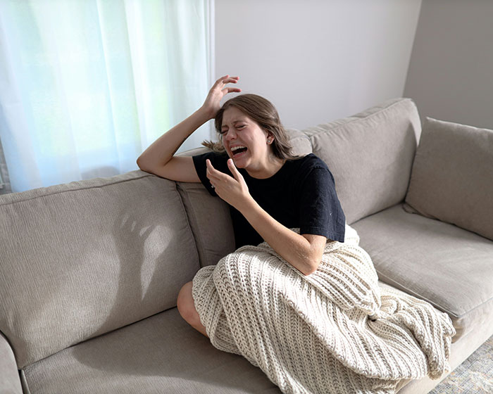 Young woman crying on couch wrapped in blanket expressing distress, relating to heavily pregnant woman stuck in bathtub scenario. Young woman crying on couch wrapped in blanket expressing distress, relating to heavily pregnant woman stuck in bathtub scenario.