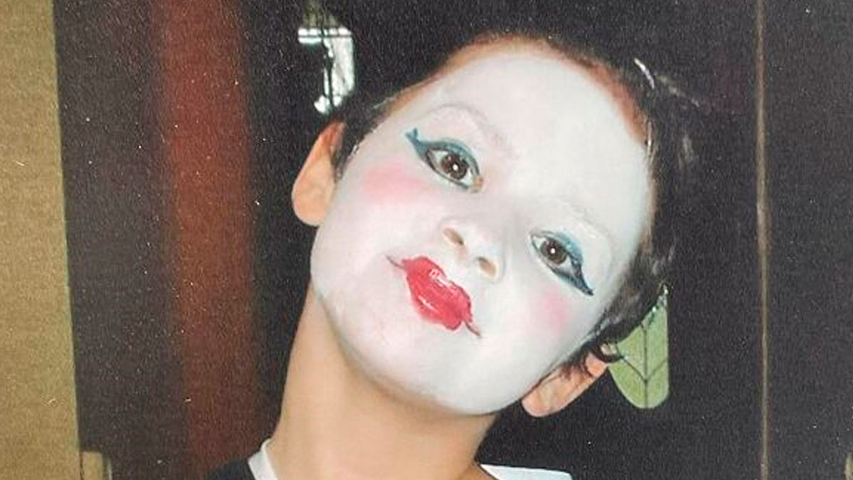 Child with regrettable past makeup, white face paint, bright red lipstick, and dramatic blue eye shadow posing for photo.