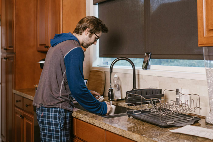 Young man washing dishes at kitchen sink, representing boyfriend ordering kind roomie to do chores like the boss.