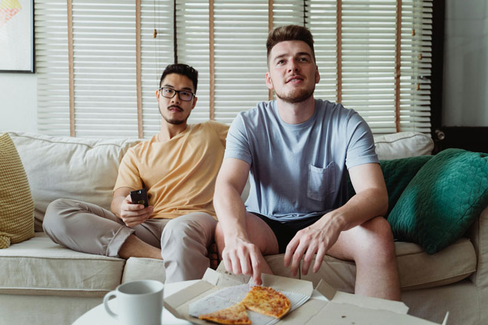 Two men sitting on a couch with pizza, capturing the dynamic of a roommate treated like the help by his bossy boyfriend.
