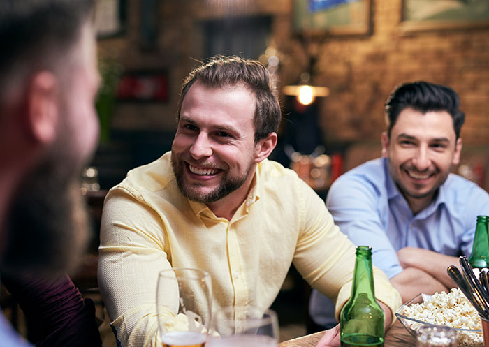 Three men smiling and chatting at a table with drinks and popcorn, illustrating a woman facing double heartbreak.