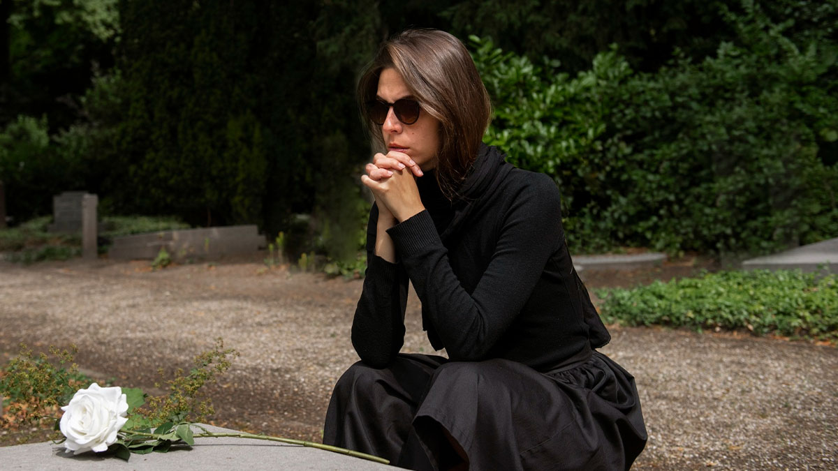 Woman in black sitting at gravesite with white rose, reflecting on widow choosing to bury late husband with first wife and child.