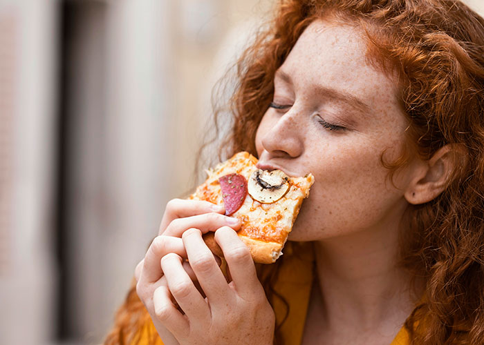 Woman enjoying a slice of pizza with mushrooms and pepperoni, highlighting a gluten-free colleague at a pizza party moment.
