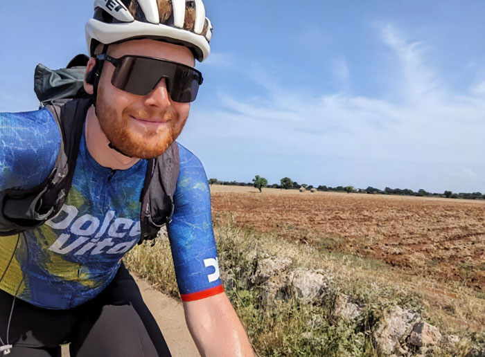 Cyclist wearing helmet and sunglasses riding along rural path with fields under clear blue sky in daytime. Cyclist wearing helmet and sunglasses riding along rural path with fields under clear blue sky in daytime.