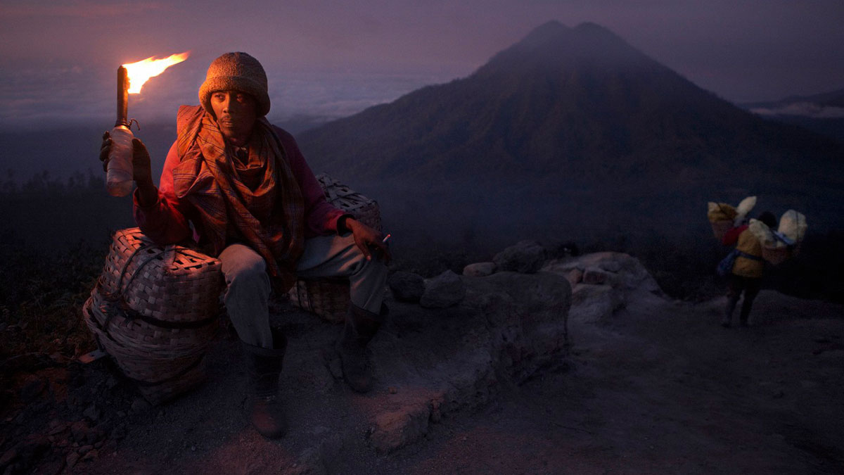 Man holding a torch sits on woven baskets in dim light with mountain in background, showcasing color photography.