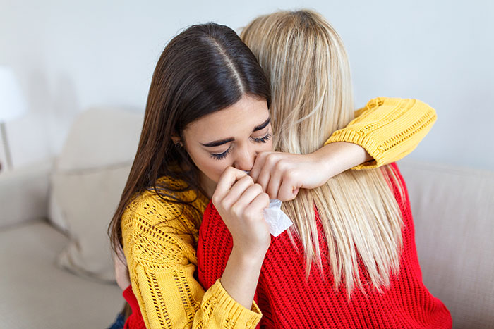 Two women hugging on a couch, one crying and holding a tissue after a racist insult involving black parents at dinner. Two women hugging on a couch, one crying and holding a tissue after a racist insult involving black parents at dinner.