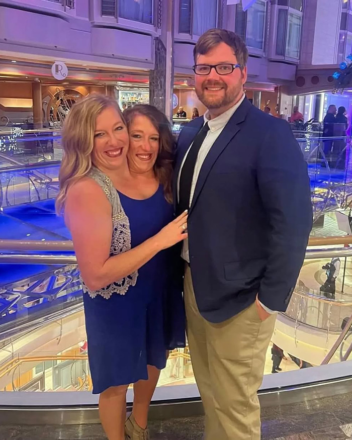 Two Hensel twins in blue dresses with a man in a suit, sparking discussion about motherhood and marriage. Two Hensel twins in blue dresses with a man in a suit, sparking discussion about motherhood and marriage.