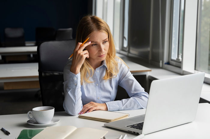 Woman in office appearing concerned during unconscious bias training, holding pencil near temple and looking at laptop screen.