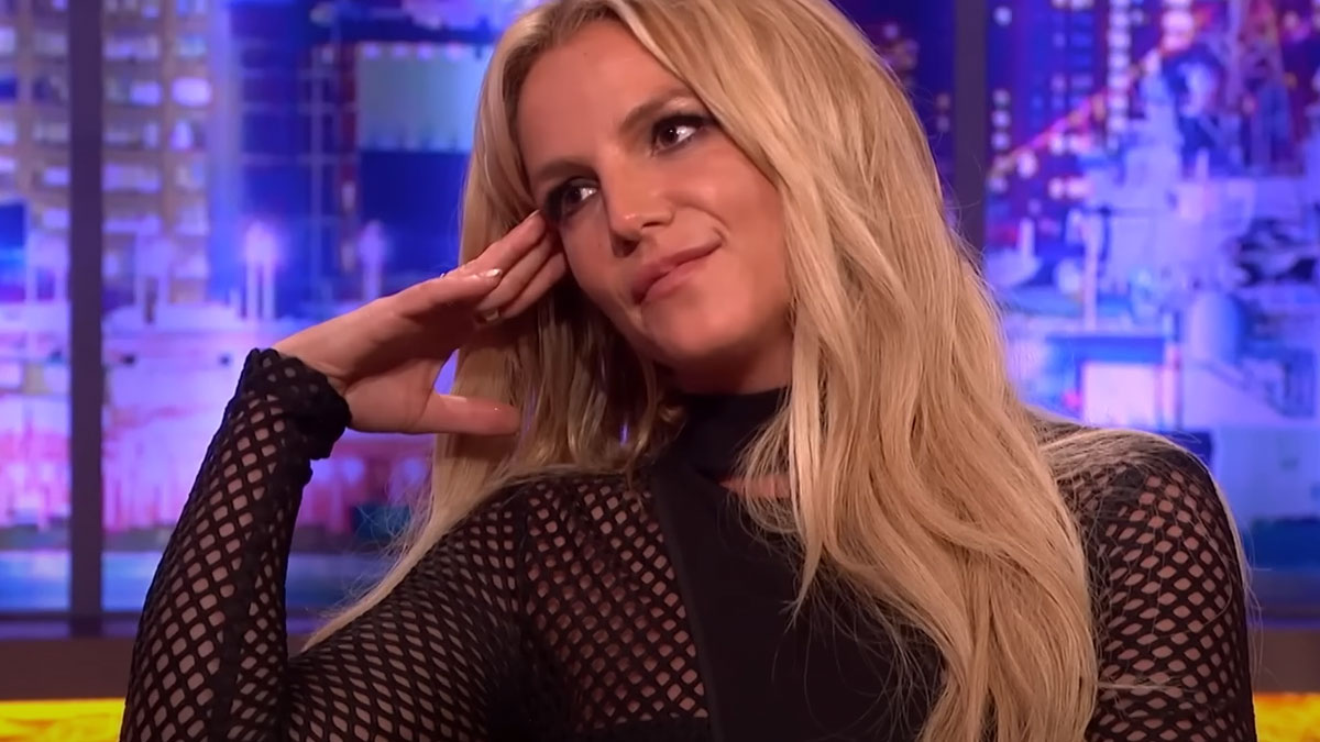Blonde woman in a black mesh top thoughtfully listening during a brightly lit indoor interview setting with cityscape backdrop.