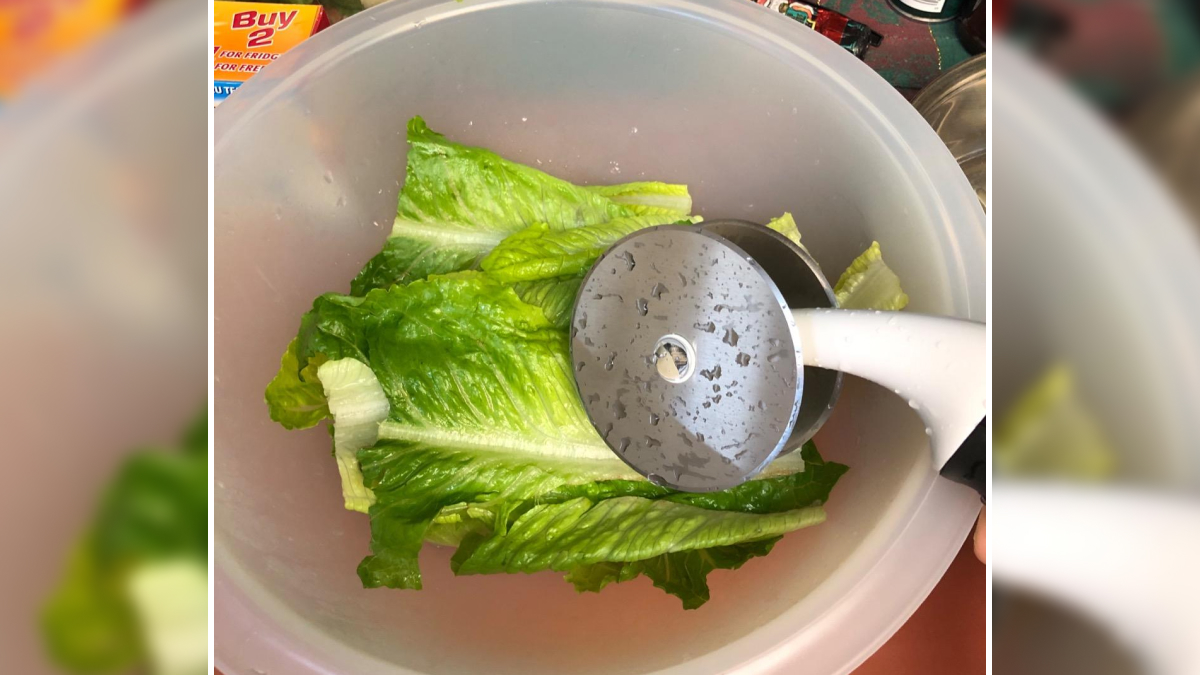 Pizza cutter being used to slice fresh green lettuce leaves in a plastic bowl among smart kitchen tools.