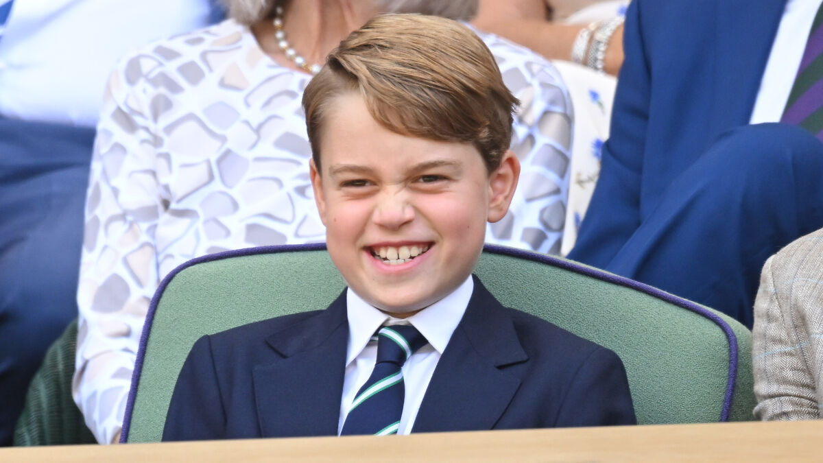 Prince George smiling in a navy blazer and striped tie, enjoying a public event with onlookers behind him.