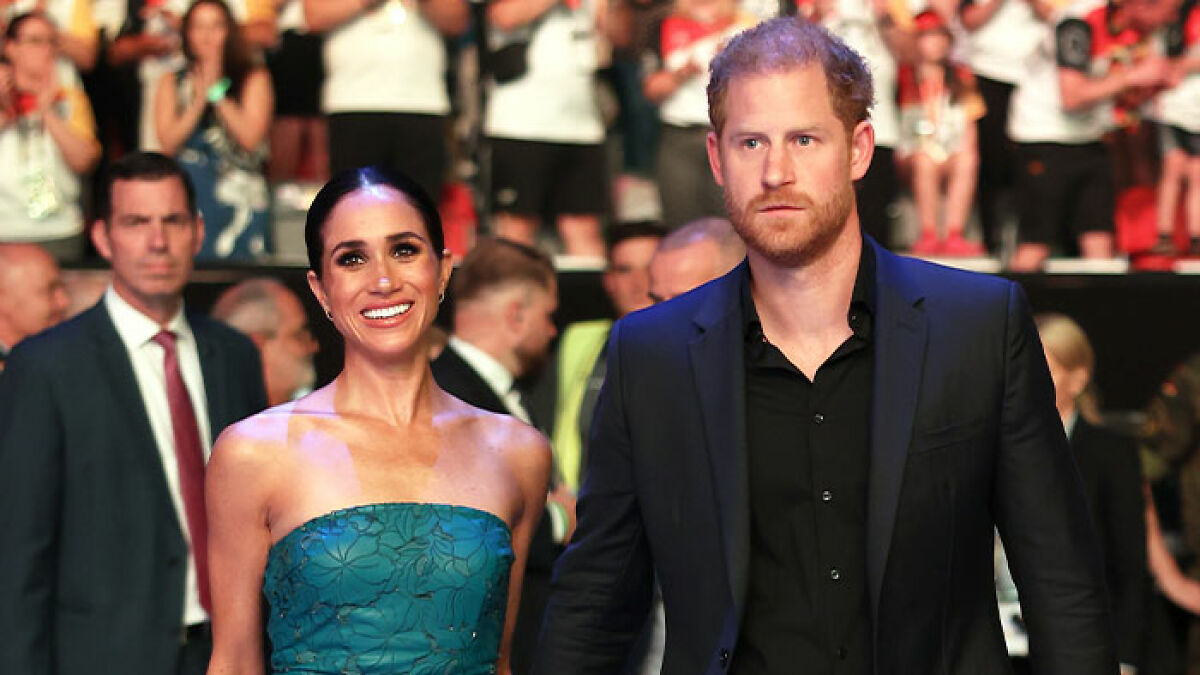 Prince Harry and Meghan Markle at a public event, dressed formally amidst a crowd in the background.