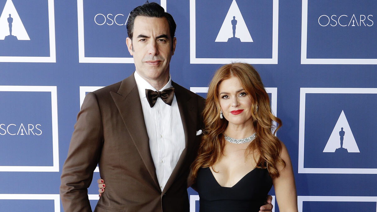 Isla Fisher and Sasha Baron Cohen posing together at an Oscars event with blue backdrop and logos.