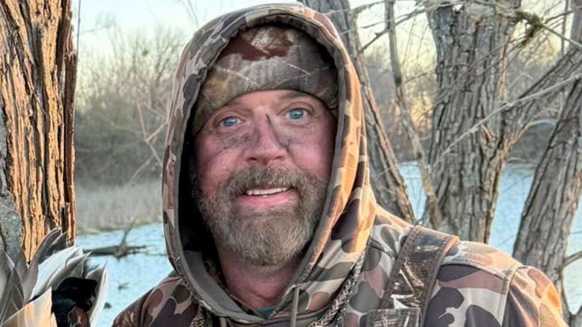 Bearded man in camouflage gear outdoors near trees and water, with face dirtied from trophy hunting activities on safari.