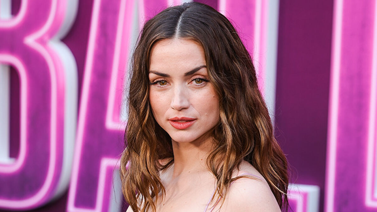 Ana de Armas at an event, pictured with wavy hair and natural makeup amid a purple-pink backdrop.