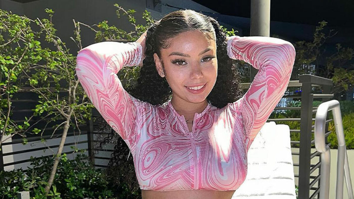 Young adult movie star posing outdoors at night, wearing a pink patterned top and smiling with hands behind her head.