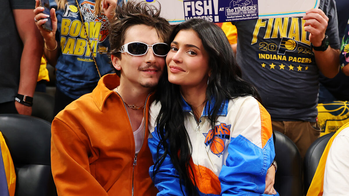 Timothu00e9e Chalamet wearing sunglasses with a woman at a basketball game, showing affection crowds around them.