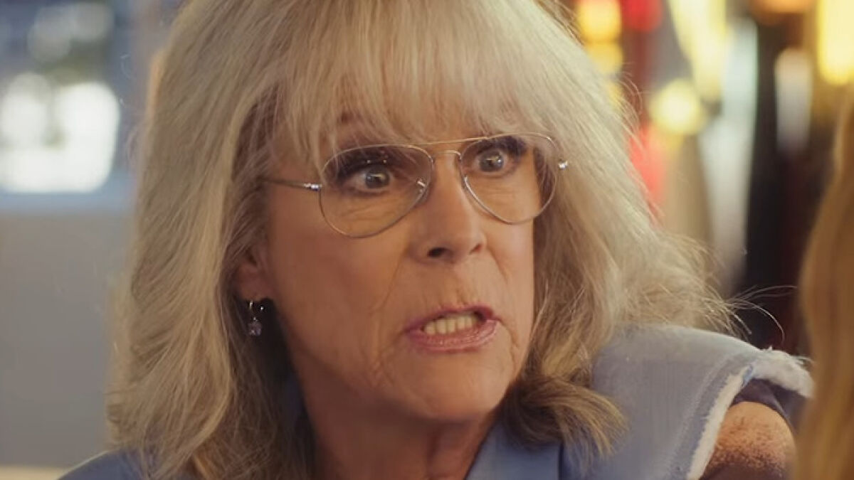Jamie Lee Curtis with dramatically fuller lips, wearing glasses and a blue top in a close-up indoor setting.