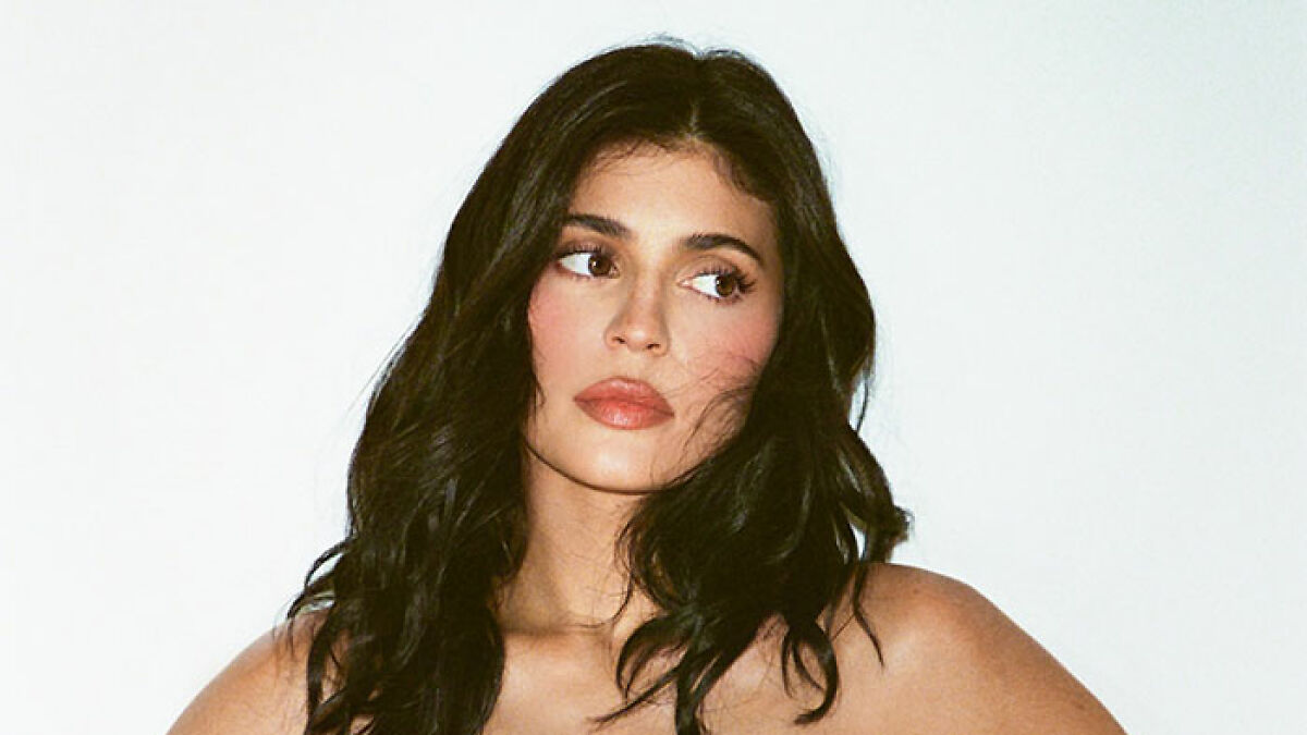 Kylie Jenner with dark wavy hair and n**e makeup posing against a white background for a racy social media post.