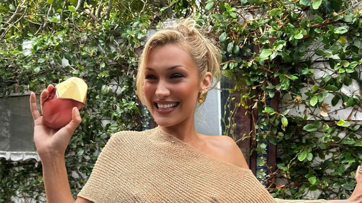 Bella Hadid smiling outdoors, holding perfume, wearing a daring braless look with a beige off-shoulder top and gold earrings.