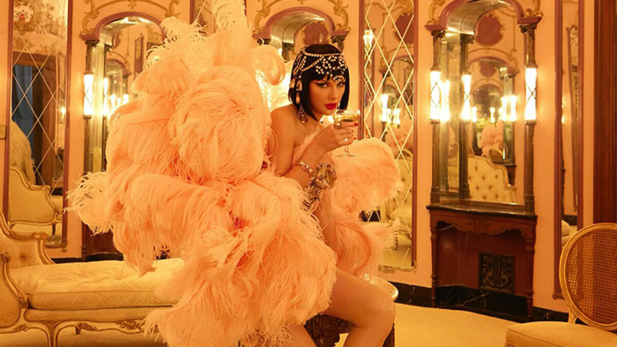 Taylor Swift in peach feathered outfit holding a glass, posing in a vintage room, sparking cover art controversy.