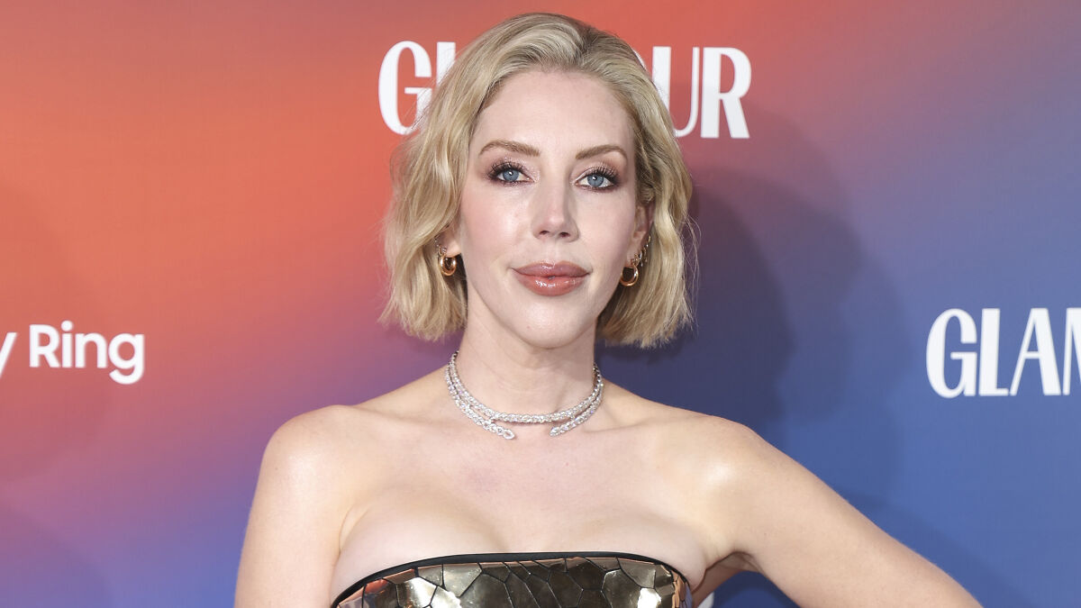 Comedian at an event, posing confidently with short blonde hair and wearing a metallic strapless dress against a colorful backdrop.