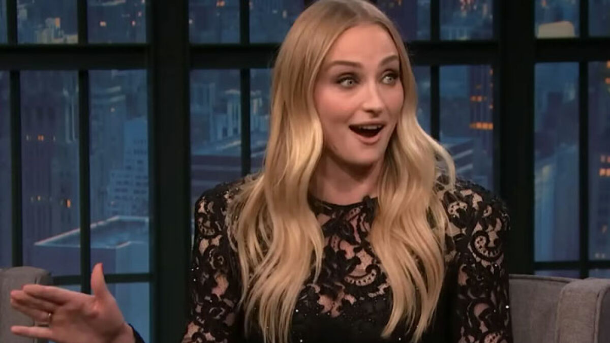 Sophie Turner speaking animatedly in a black lace dress during a late night talk show interview.