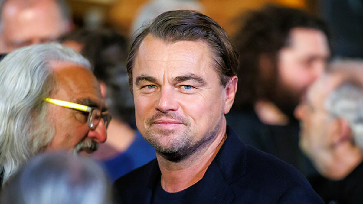 Leonardo DiCaprio smiling in a crowded event, fans discussing his PDA-filled yacht trip with girlfriend.