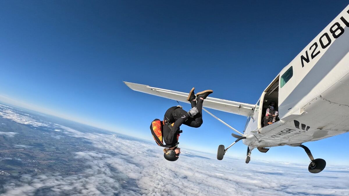 Woman skydiving from 15,500-ft altitude after breakup, captured mid-flip beside small airplane in clear blue sky.