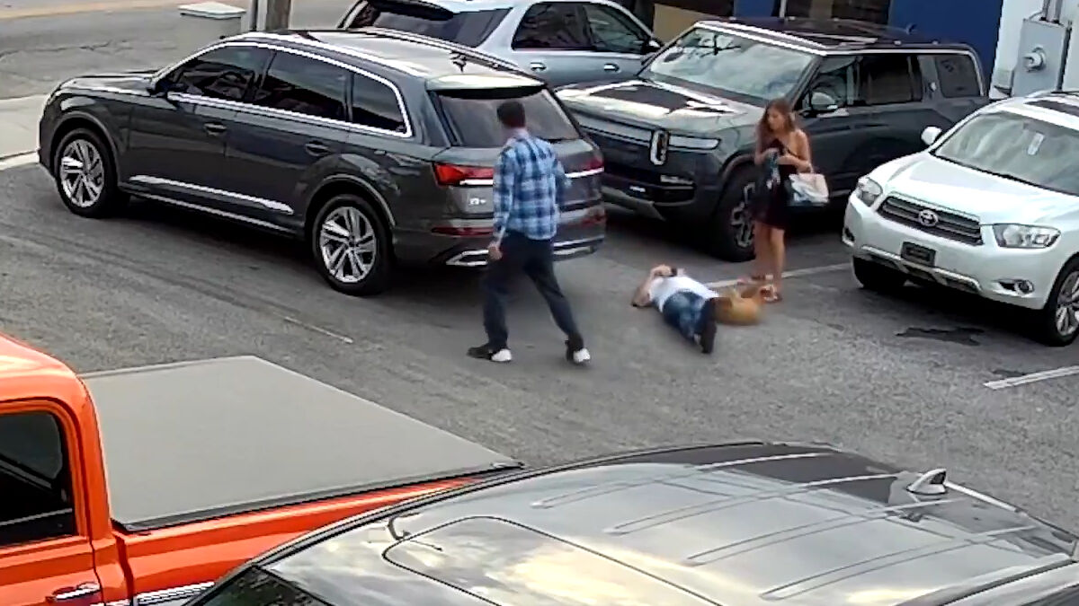 Man lies on the ground next to a service dog, with an Uber driver and a woman standing nearby in a parking lot.
