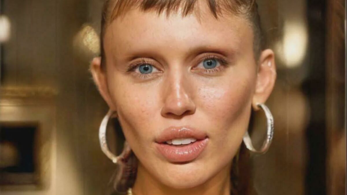 Close-up of a person with no eyebrows, blue eyes, and hoop earrings highlighting the trend of eyebrow-free fashion.
