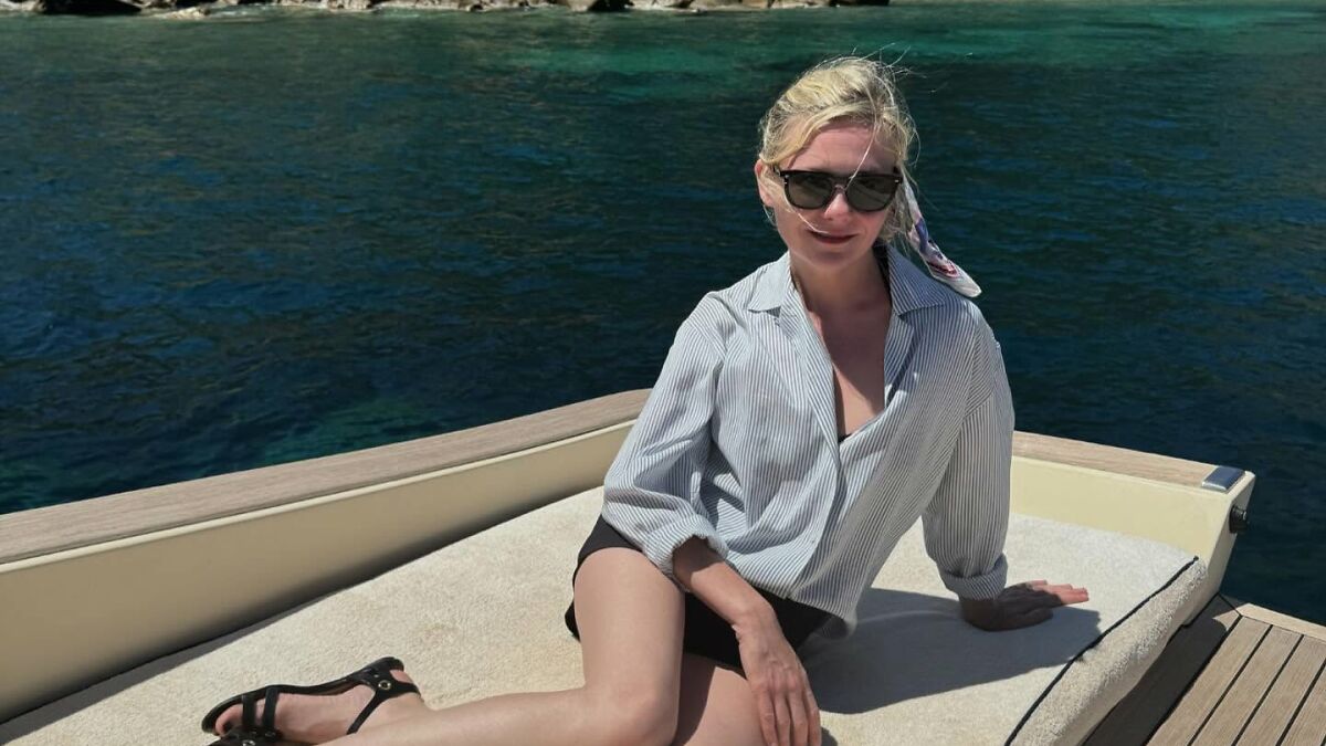 Kirsten Dunst sitting on a boat with sunglasses and a striped shirt, enjoying a sunny day by the water.