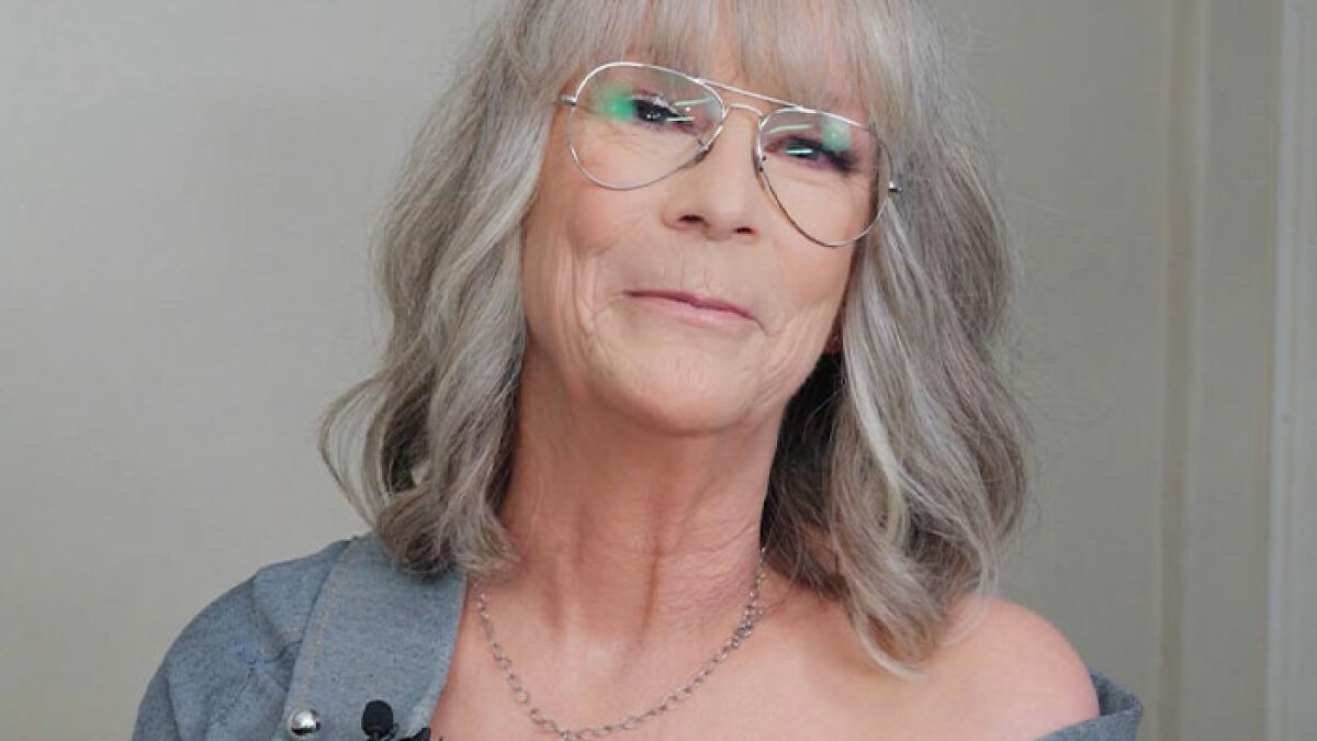 Jamie Lee Curtis smiling, wearing glasses and a gray top, in a casual setting responding to fan memes on viral cleavage moment.