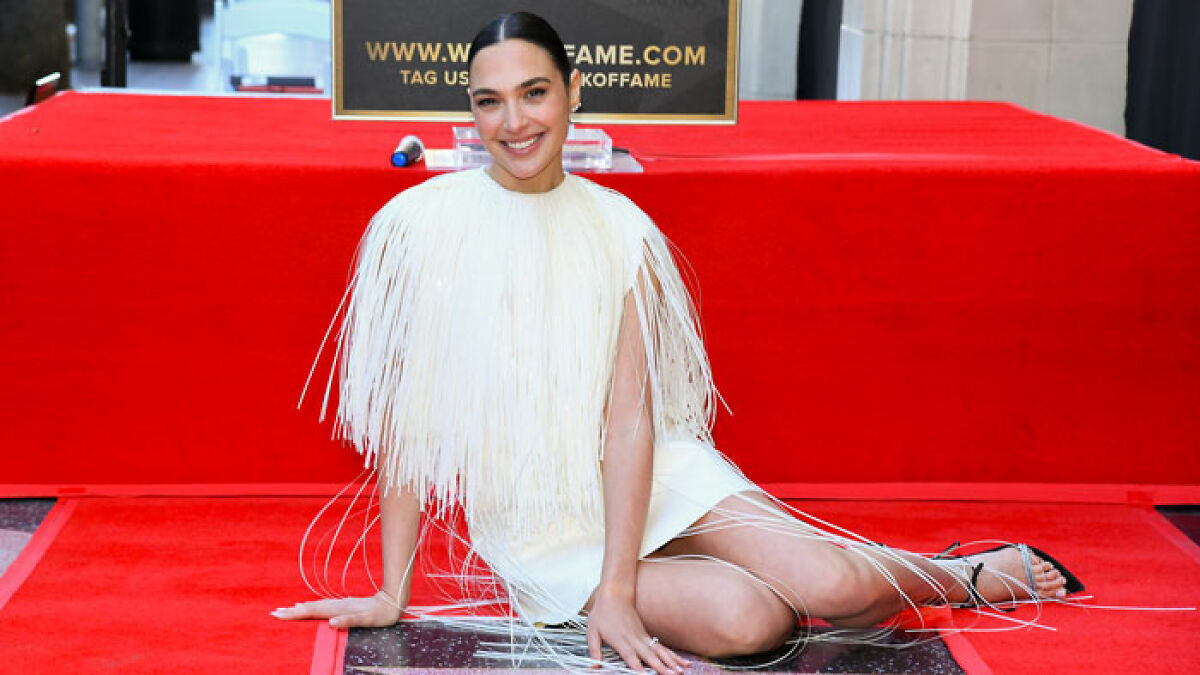 Gal Gadot wearing a white fringed dress, smiling while seated on a red carpet at a Hollywood event.