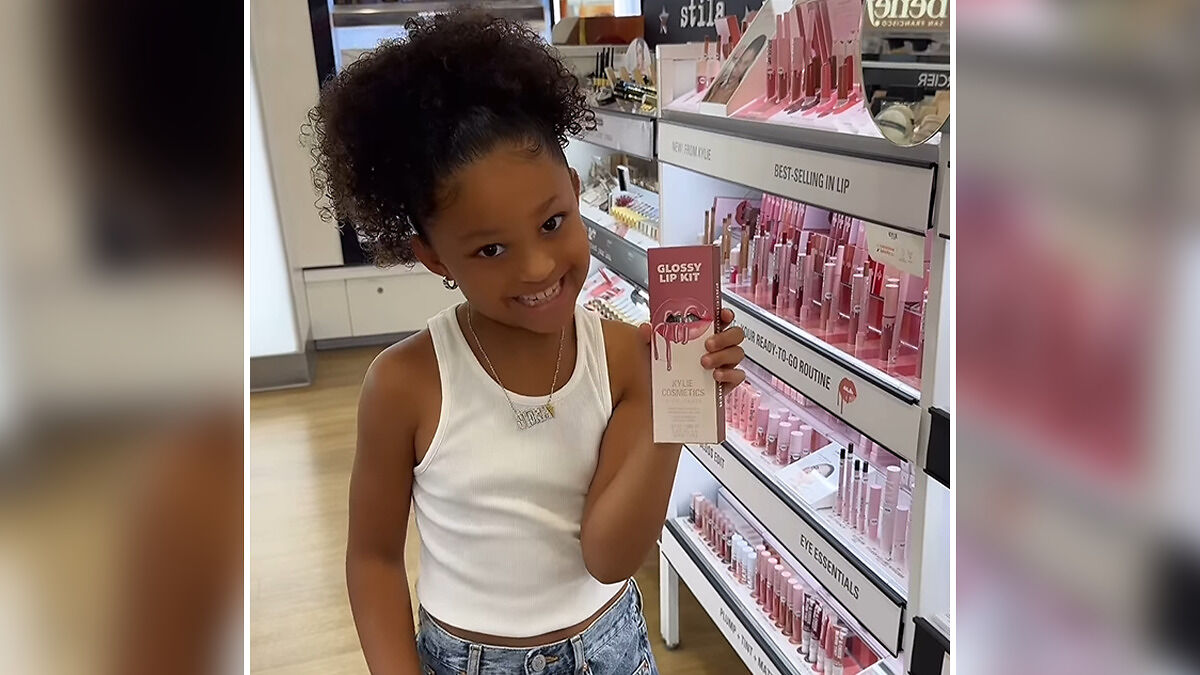 Stormi Jenner holding Kylie Cosmetics Glossy Lip Kit smiling in a store with makeup display in the background