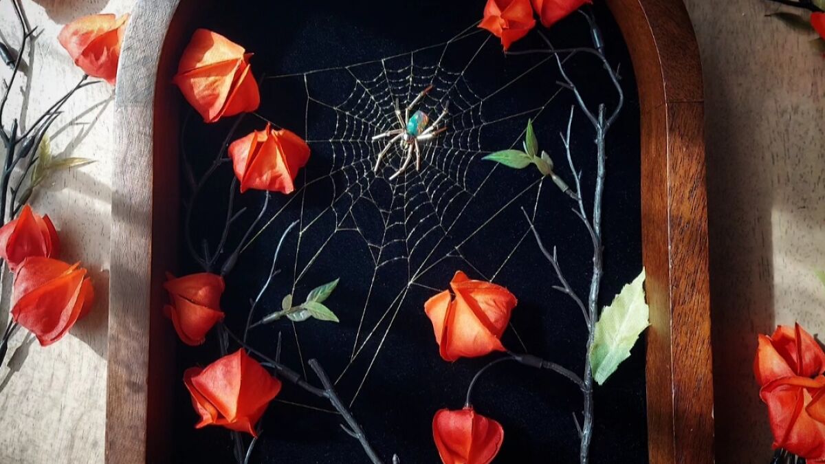 Handmade spider web art displayed with red flowers and branches inside a wooden frame on a beige surface.