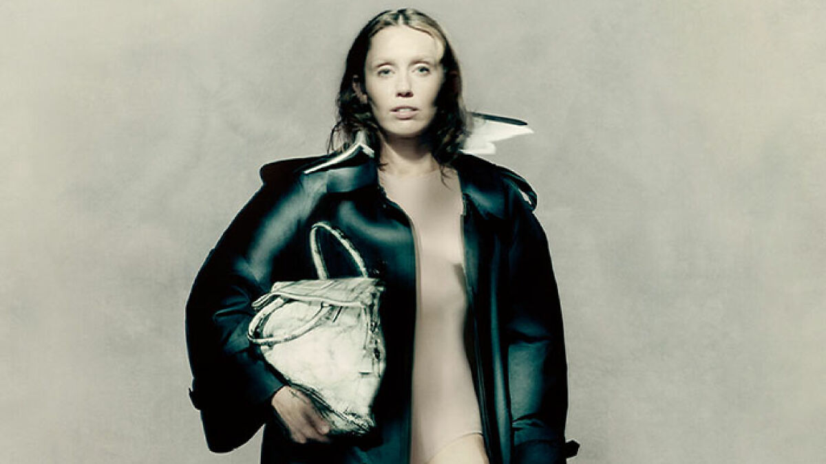 Model wearing body paint and oversized black jacket holding a large bag for Maison Margiela campaign.