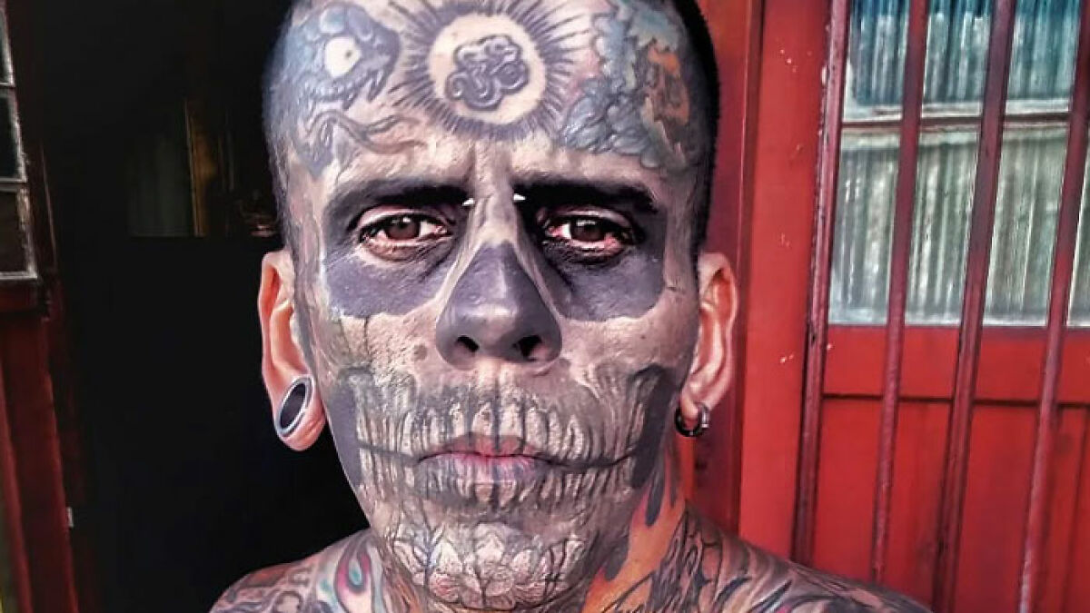 95% tattooed Brazilian Steve-O man showing dramatic new look after tattoo removal standing by red door.