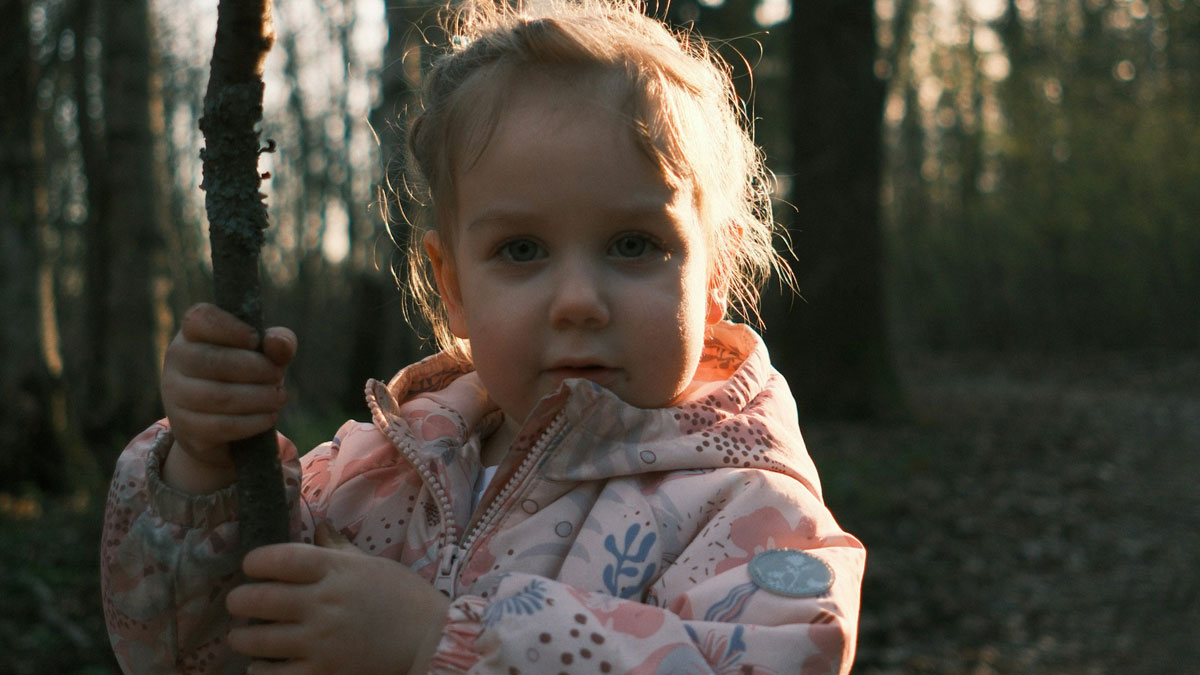Young girl in a forest holding a stick with sunlight highlighting her face, evoking creepy and terrifying imaginary friends.
