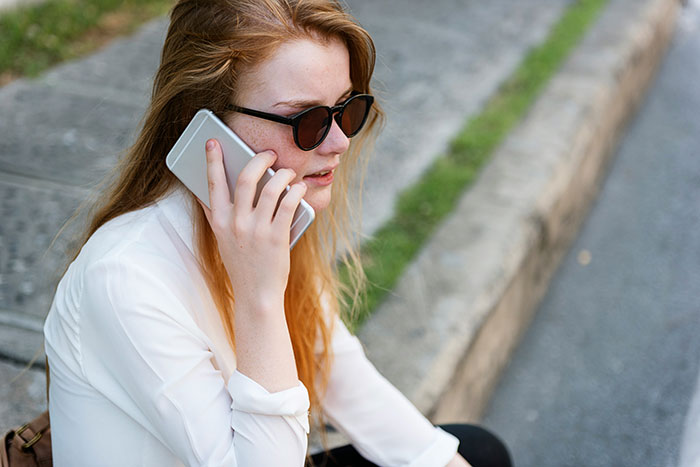 Young woman wearing sunglasses sitting outdoors, looking concerned while talking on her phone about lazy parenting issues.