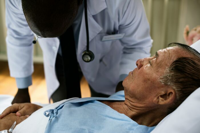 Doctor in white coat with stethoscope leaning over and talking to elderly male patient in hospital bed, illustrating gender-based double standards.