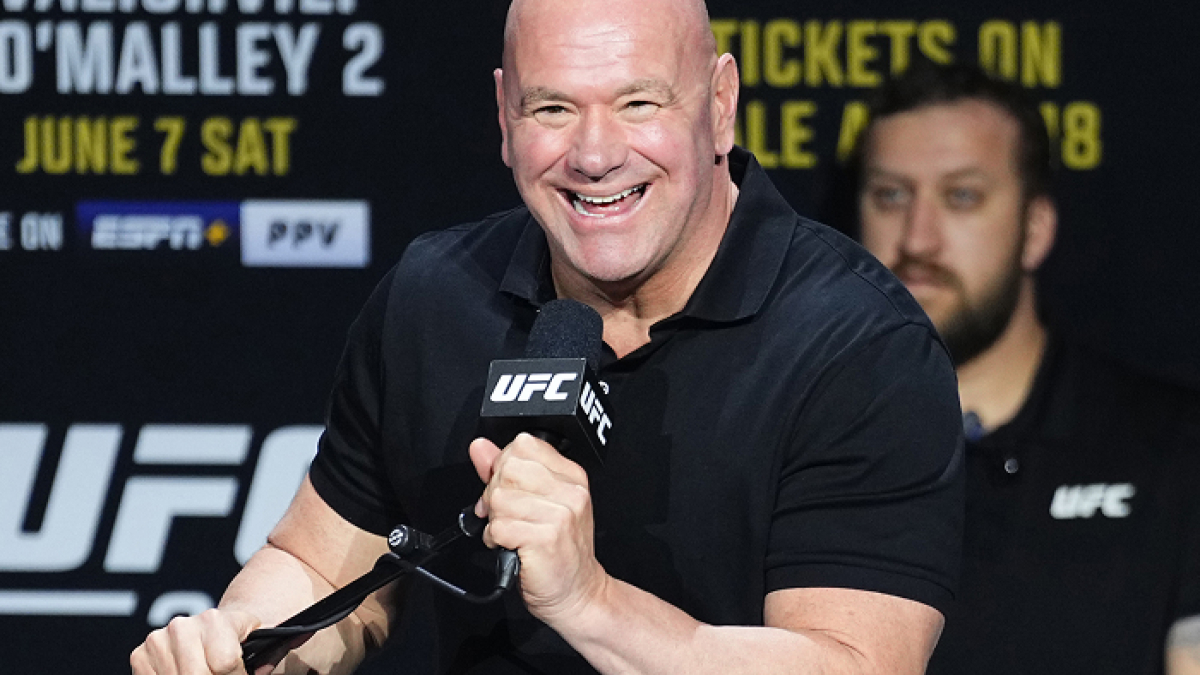 Dana White speaking at UFC event press conference, smiling and holding a microphone with UFC branding visible.
