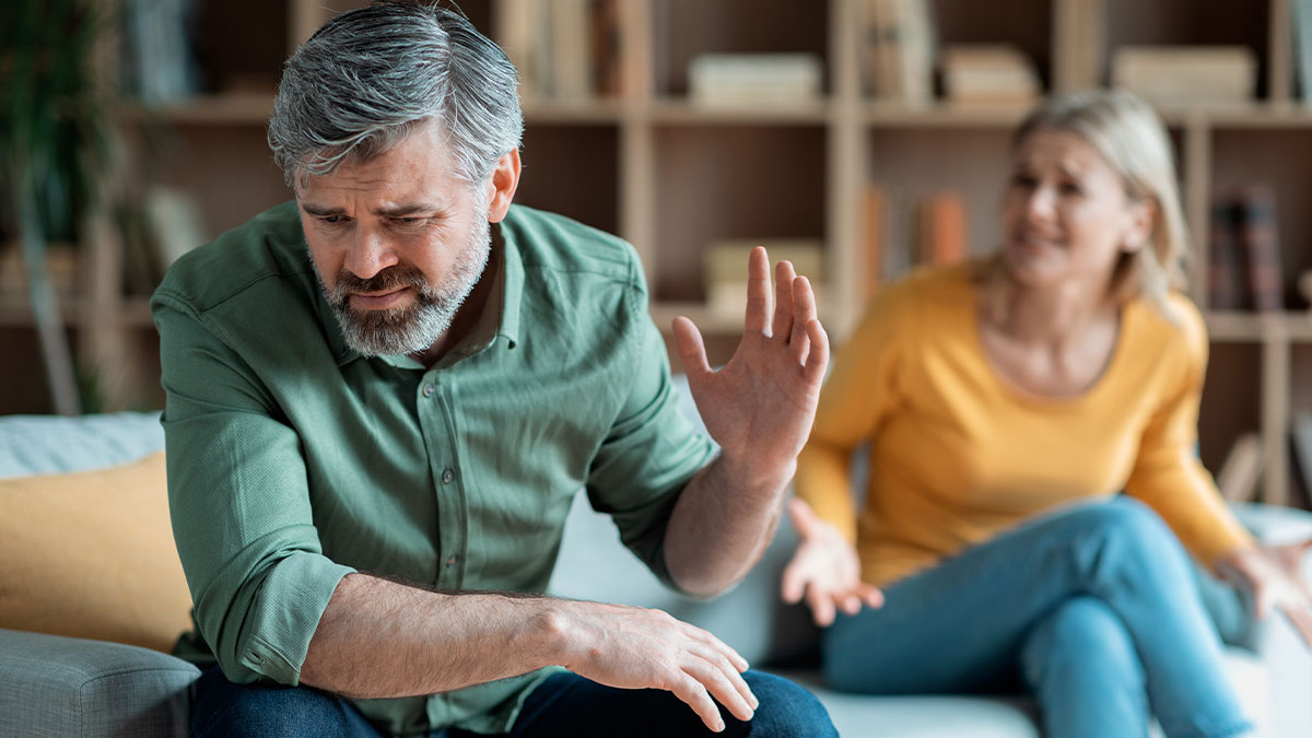 Man with gray hair and beard upset in living room while woman in yellow argues behind him, reflecting cheating texts tension.