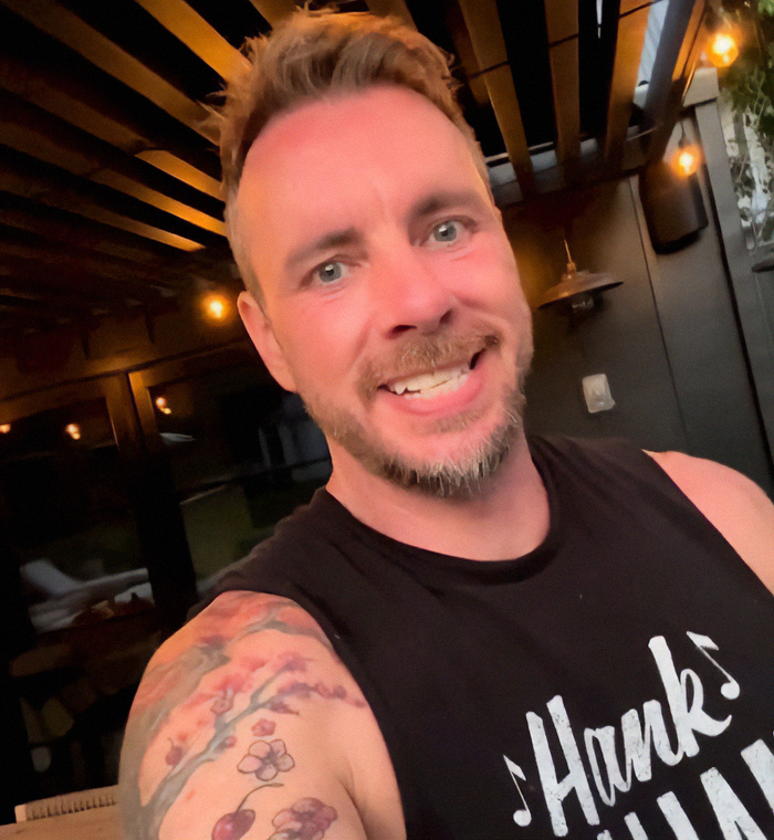 Dax Shepard smiling in a black tank top with a floral tattoo on his arm in a warmly lit indoor setting. Dax Shepard smiling in a black tank top with a floral tattoo on his arm in a warmly lit indoor setting.