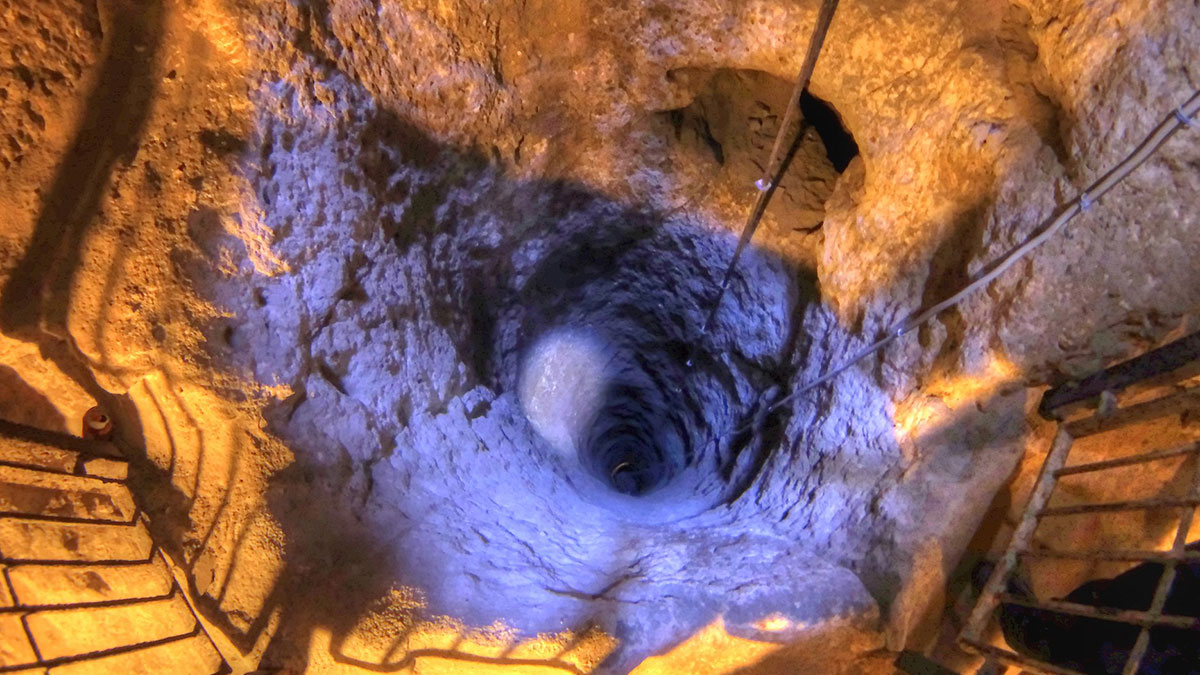 Deep vertical shaft inside a mysterious underground city, showing rugged stone walls and metal ladders with artificial lighting.