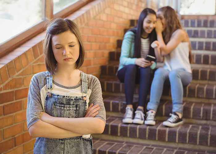 Teen girl with crossed arms looking upset while two friends whisper behind her on brick stairs, reflecting relationship issues.