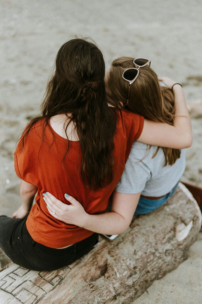Two friends sitting on a log at the beach, one with sunglasses on her head, highlighting friendship and trust before a shocking discovery.
